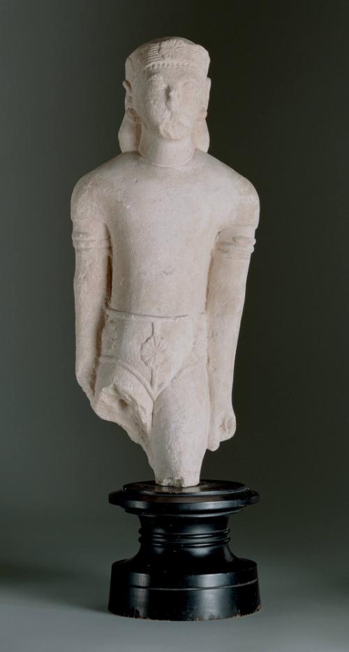 Male statuette wearing short-sleeved tunic, shorts with rosette flowers, rosette flower headband, spiral earrings, and snake-headed spiral arm bracelets