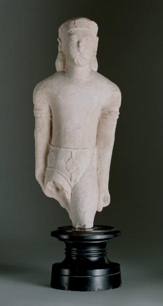 Male statuette wearing short-sleeved tunic, shorts with rosette flowers, rosette flower headband, spiral earrings, and snake-headed spiral arm bracelets