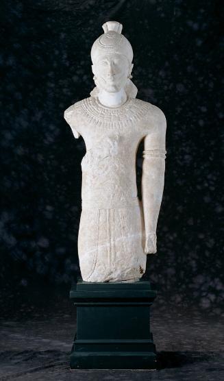 Male statue wearing a tunic, kilt with uraeus snakes on the sash, lotus-flower-knobbed cap with a rosette flower and earflaps folded up, chin strap down cheeks and under chin, spiral earrings, spiral arm bracelet with rosette flower, and three-tiered pectoral necklace