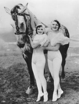 Two female performers from the Loyal-Repenski Troupe stand beside a horse each with one arm on …