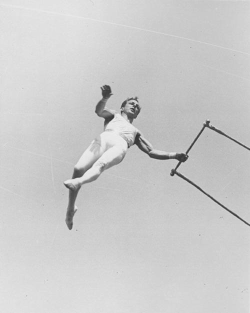 Alfredo Cordona swinging from a trapeze with his left hand on the trapeze bar.