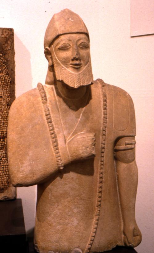Male statue with full beard wearing a tunic, fringed cloak, conical cap with ear flaps folded up, and spiral arm bracelet