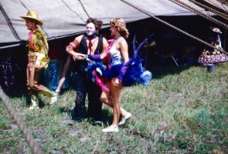 2 men in costumes (cowboy and clown) and a lady in purple, pink, and silvery costume