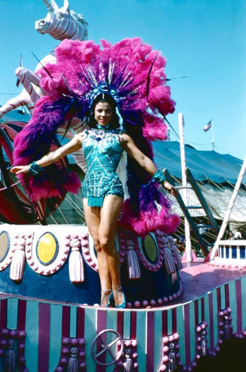 Lady in teal costume with purple/pink headress in front of a unicorn float